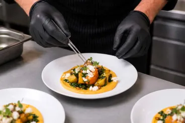 A person wearing gloves carefully plating a gourmet dish with roasted vegetables, greens, and cheese on a white plate in a kitchen setting.