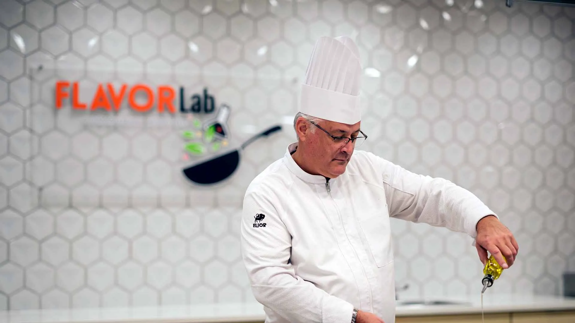 A chef in a white uniform and tall hat is pouring oil into a pan while cooking in a modern kitchen labeled Flavor Lab.