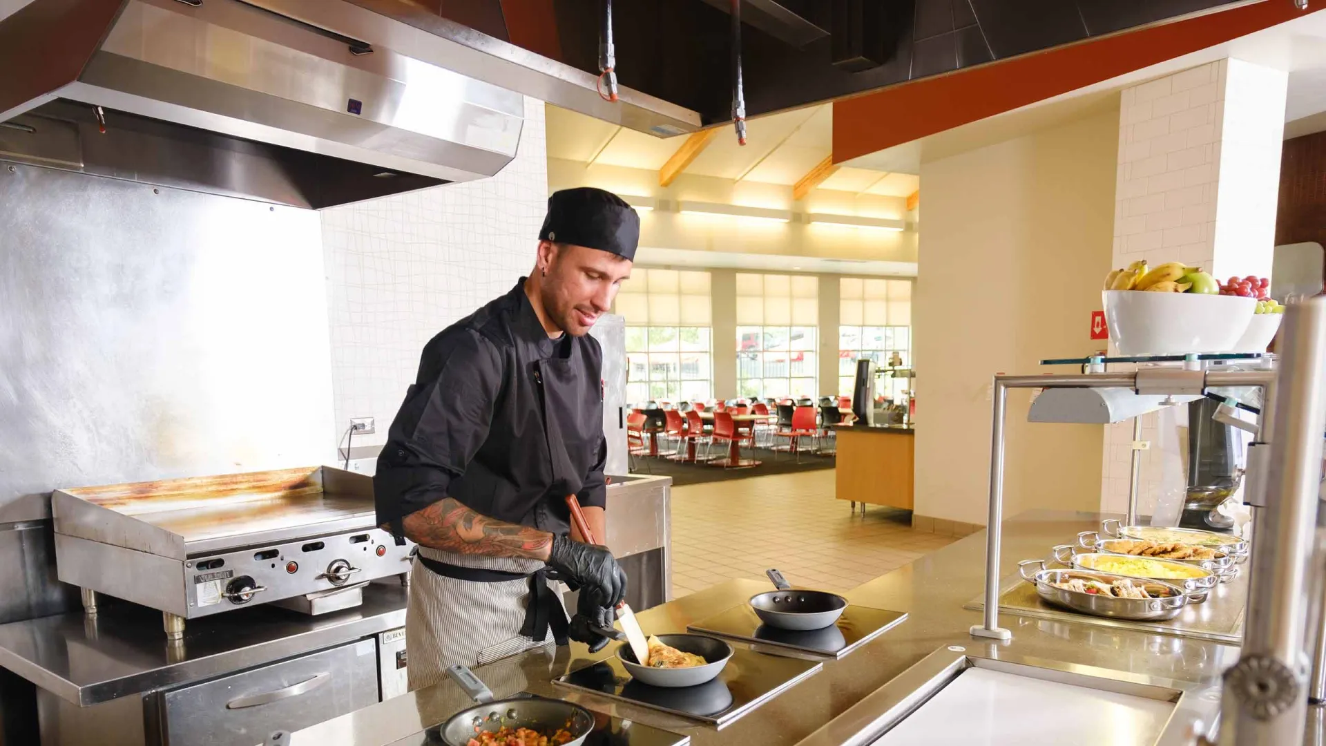 A chef wearing a black uniform and gloves is cooking and stirring food in pans on a stovetop inside a clean, well-lit commercial kitchen.