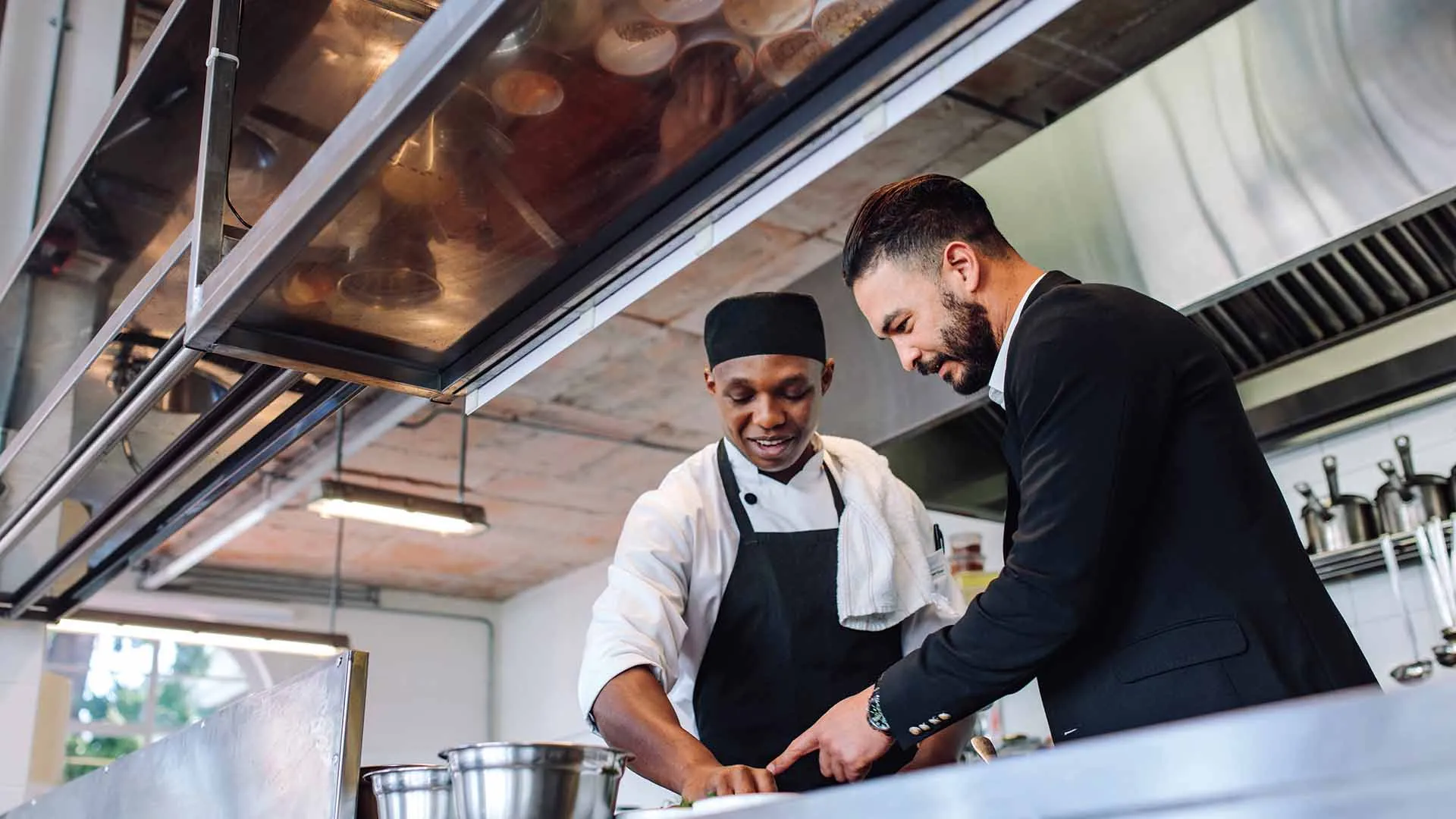 A chef and a man in a suit collaborating and discussing something over a kitchen counter in a professional restaurant kitchen.