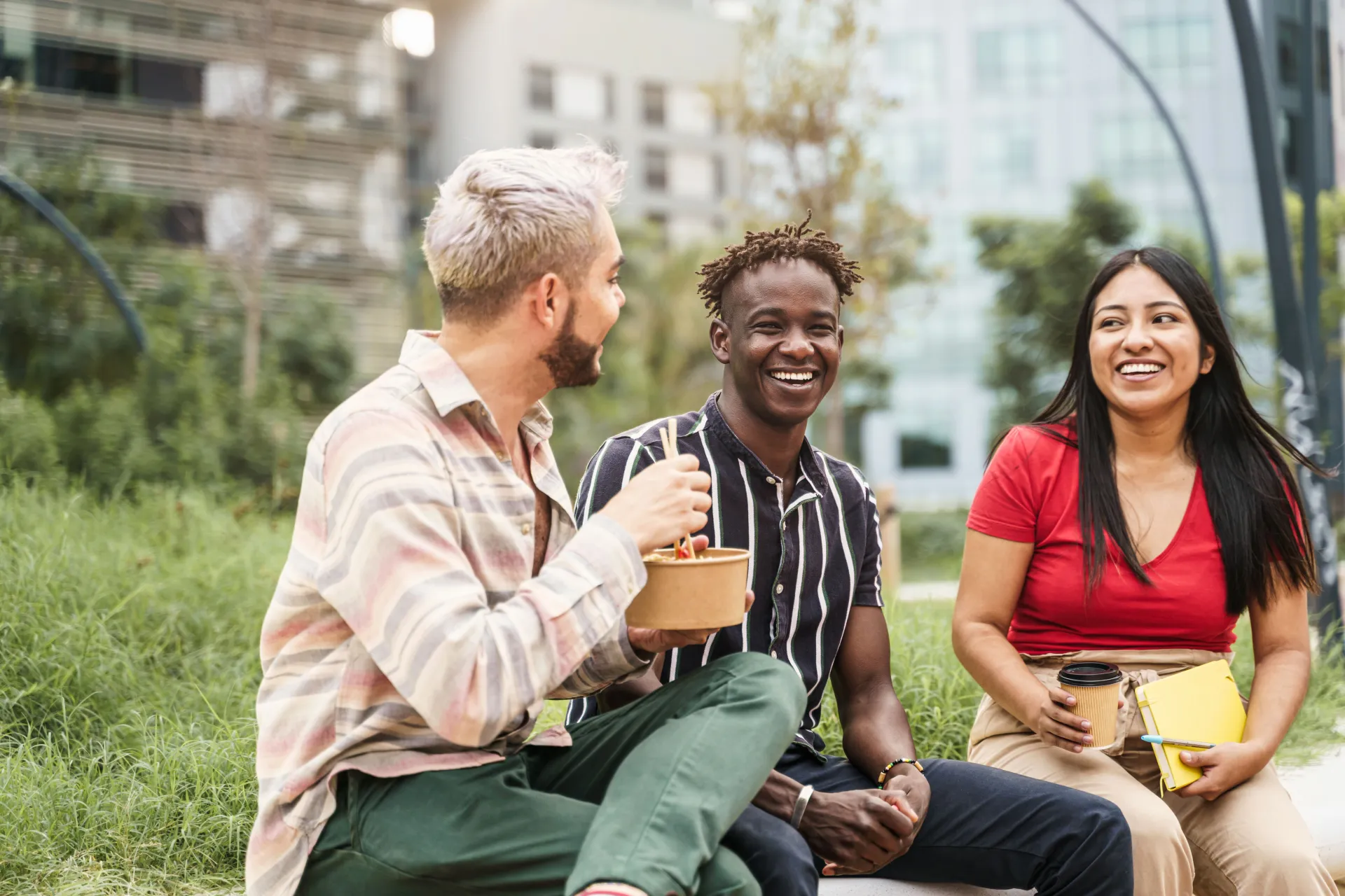 Three young adults are sitting on the grass in an urban park, smiling and enjoying food and drinks together.