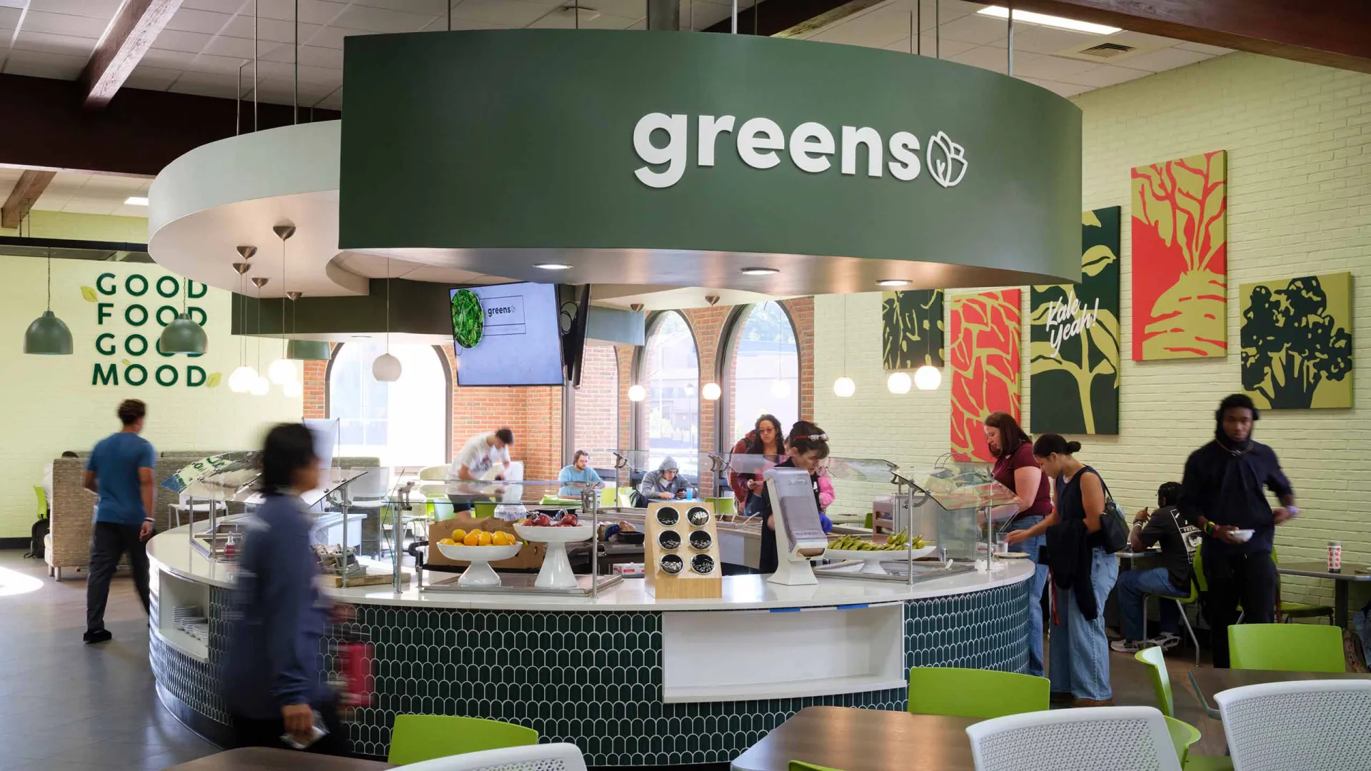 A modern cafeteria setting with people serving themselves at a circular salad bar labeled "greens," surrounded by tables and chairs with colorful, abstract wall art in the background.