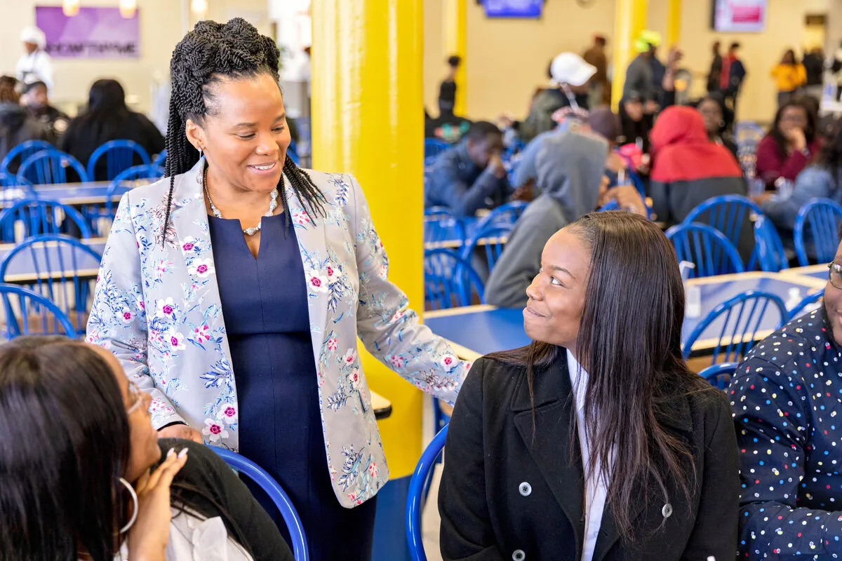 A woman standing and smiling while engaging in conversation with a seated woman in a busy cafeteria filled with people.