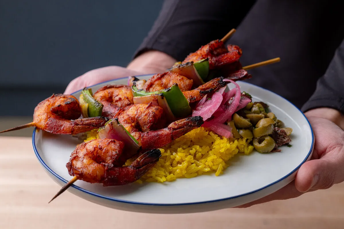 Hands holding plate of shrimp skewers, yellow rice and olives