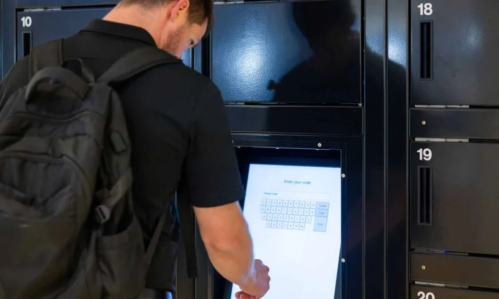 A man with a backpack is interacting with a touchscreen keypad on a black electronic locker system, likely to enter a code for access.