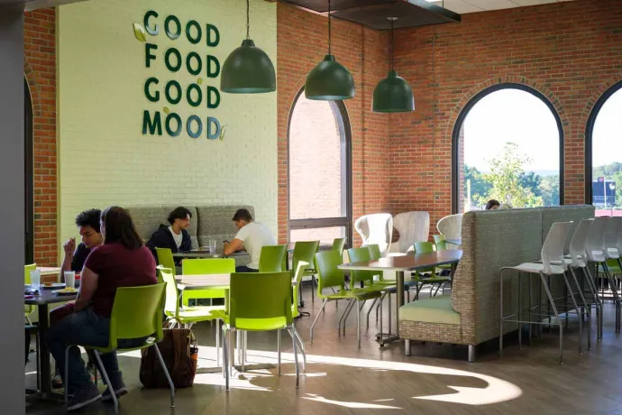 A bright, modern café with green chairs and hanging pendant lights, where people are seated and engaged in conversation or working on laptops, under a wall sign that reads "GOOD FOOD GOOD MOOD."
