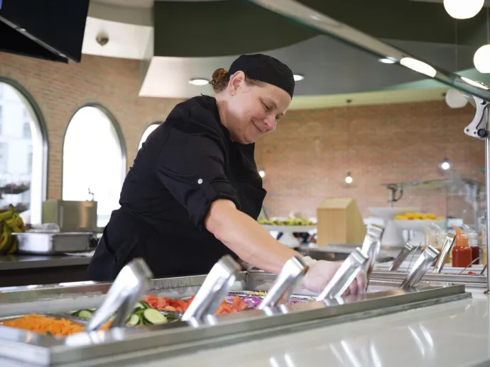 A chef wearing a black uniform and hat is serving food from a salad bar inside a cafeteria.