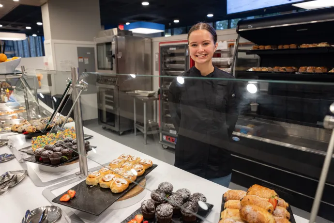 A smiling female chef stands behind a counter displaying a variety of pastries in a food service area.