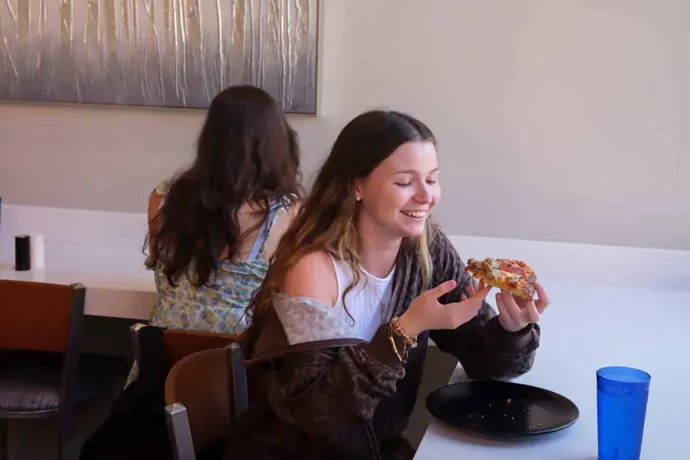 Young woman sitting at a table, eating a slice of sausage and pepperoni pizza.