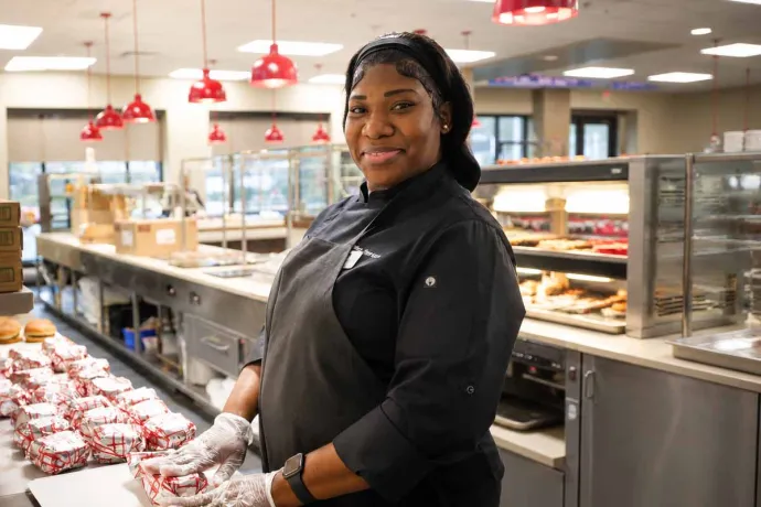 A smiling chef in a black uniform and gloves, standing behind a counter in a cafeteria, wrapping sandwiches.